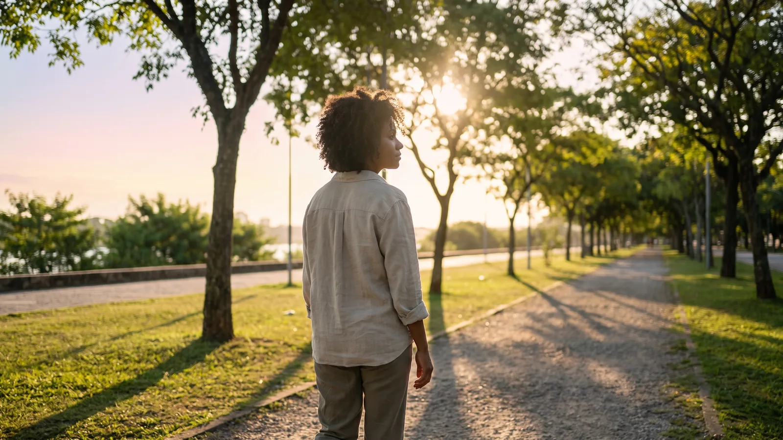 Mulher caminhando de costas por um parque verde no fim da tarde, em ritmo tranquilo e sem pressa
